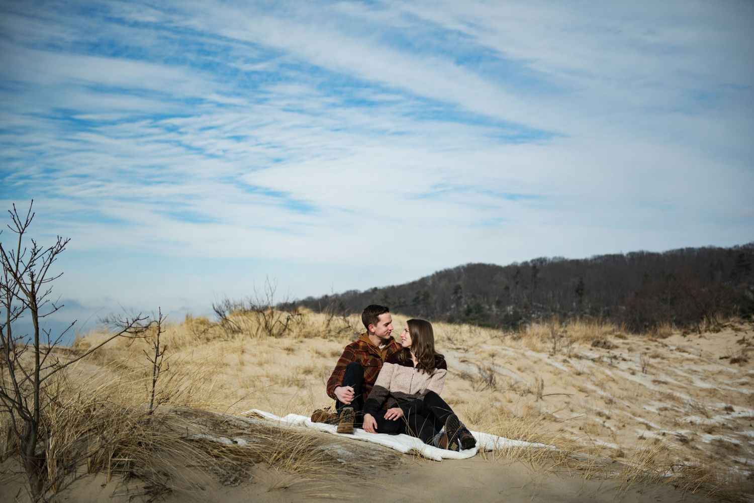 Two people sit together on a blanket overlooking a scenic winter landscape with wispy clouds in the blue sky above.