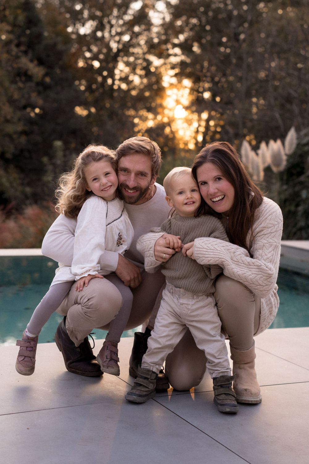 Glückliche Familie hockt in aufeinander abgestimmten creme- und beigefarbenen Outfits zusammen am Poolrand.