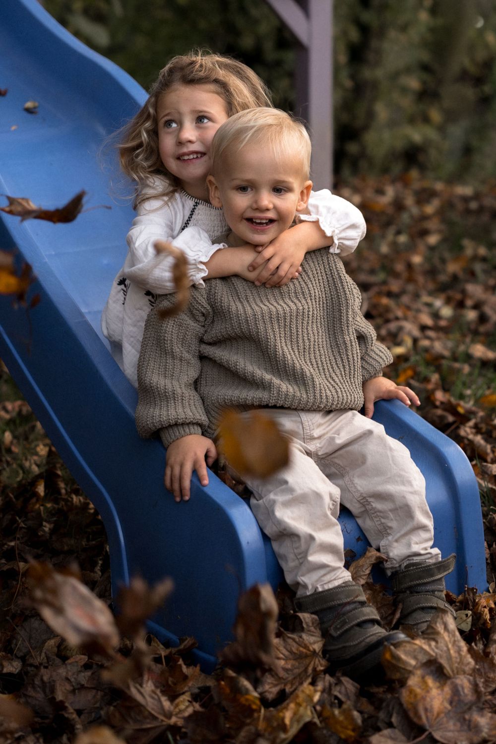 Zwei Kinder spielen zusammen auf einer blauen Spielplatzrutsche, umgeben von Herbstblättern auf dem Boden.