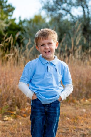 Young boy in light blue polo shirt smiles brightly while standing in an outdoor setting.