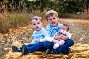 Three young siblings sit together on a golden blanket surrounded by fall leaves.