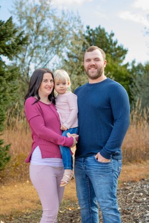 A family of three poses outdoors against a natural backdrop of trees and tall grasses.