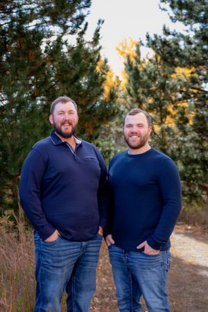 Two men in blue sweaters smile while standing on nature trail with pine trees behind them.