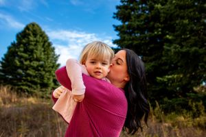 Parent giving child a loving kiss outdoors with evergreen trees in background.