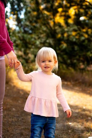 Young child in pink dress holds parent's hand while walking on autumn trail.