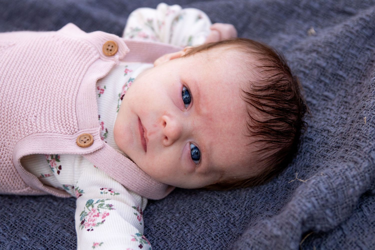 Baby wearing pink clothing lies on grey blanket looking at camera.