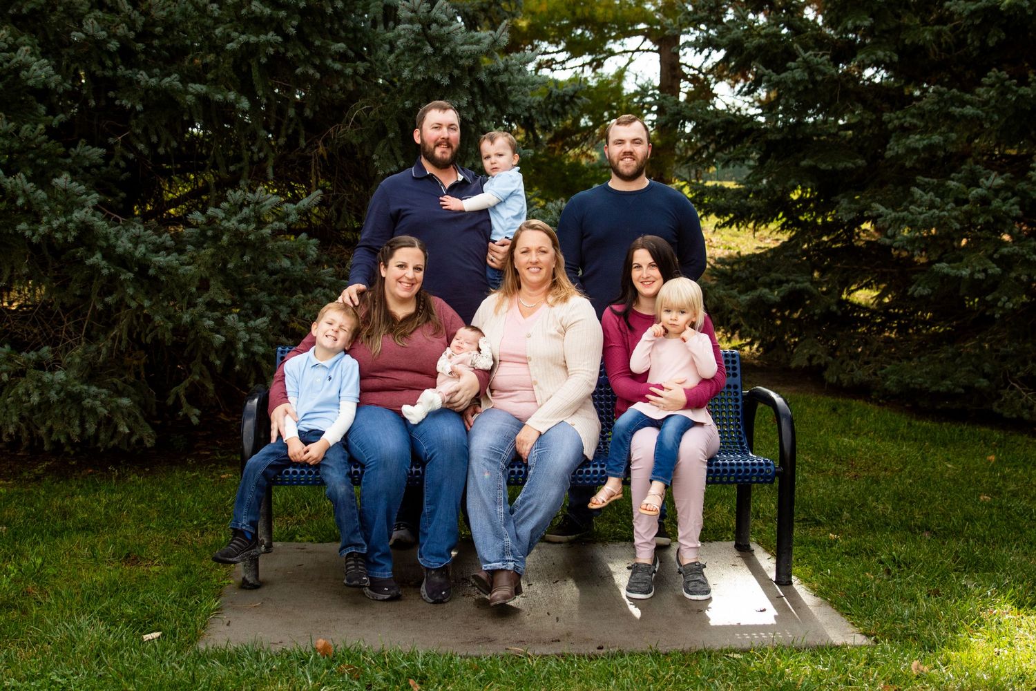 Family group sits together on a park bench surrounded by evergreen trees.