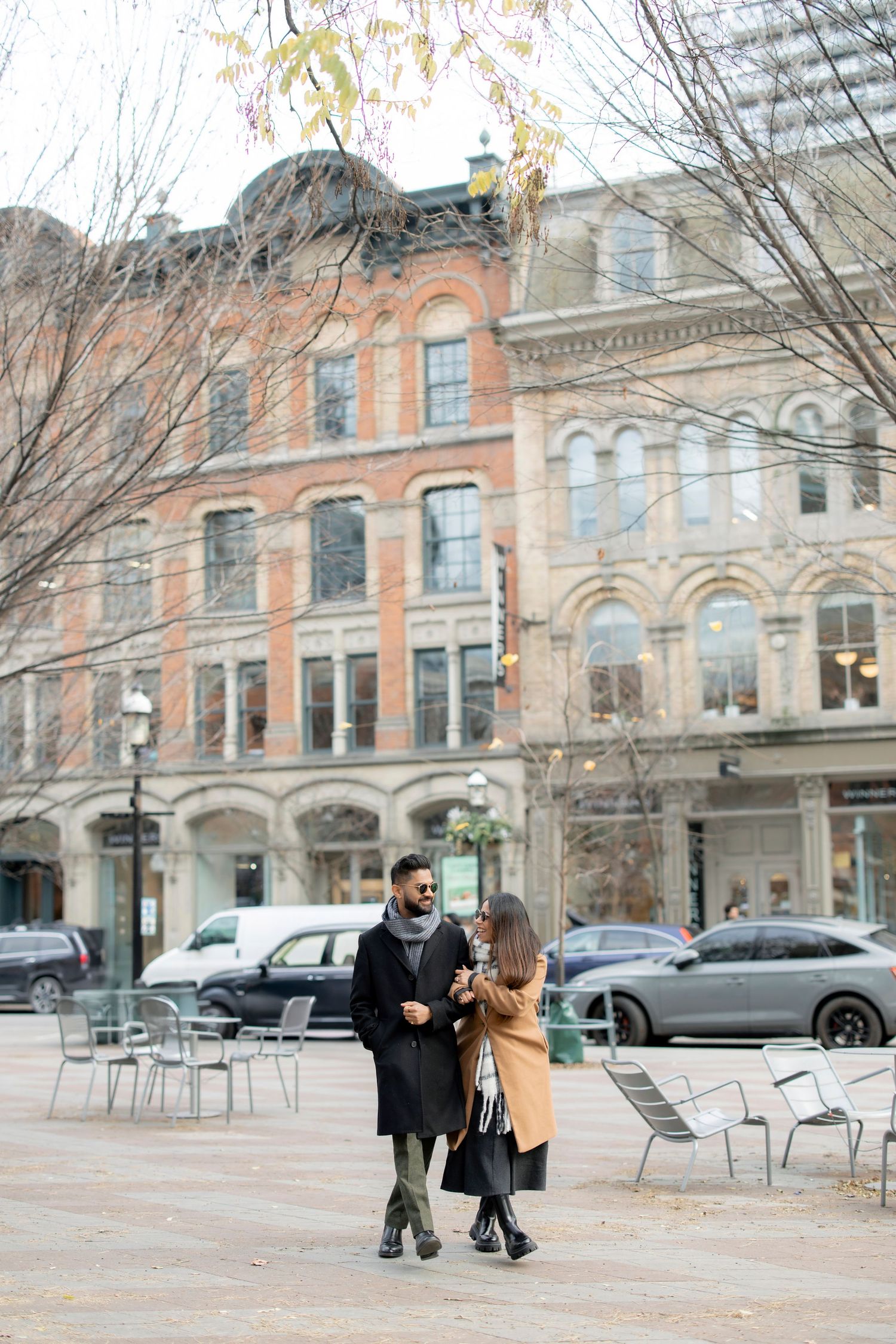 A stylish couple walking together in front of historic brick buildings on an autumn day.