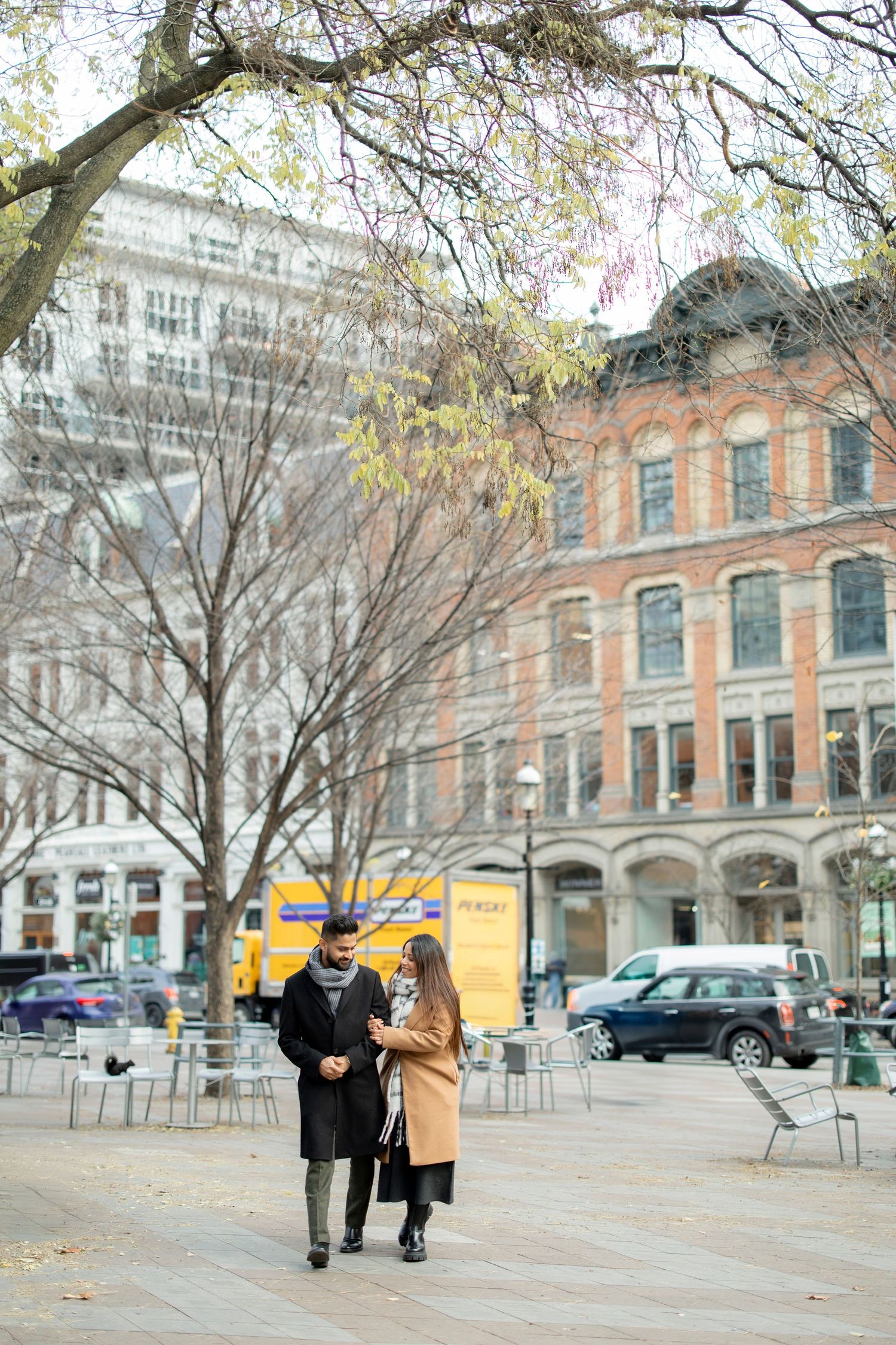 A couple walking through a plaza with historic architecture and bare autumn trees.
