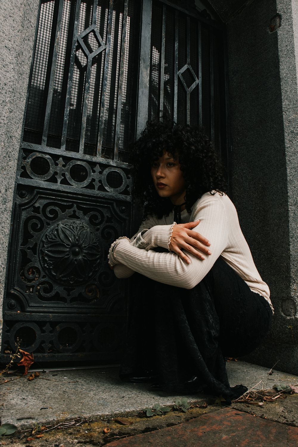 A woman with curly dark hair crouches in a stone doorway beside an ornate black wrought-iron gate, wearing a cream sweater and black skirt.