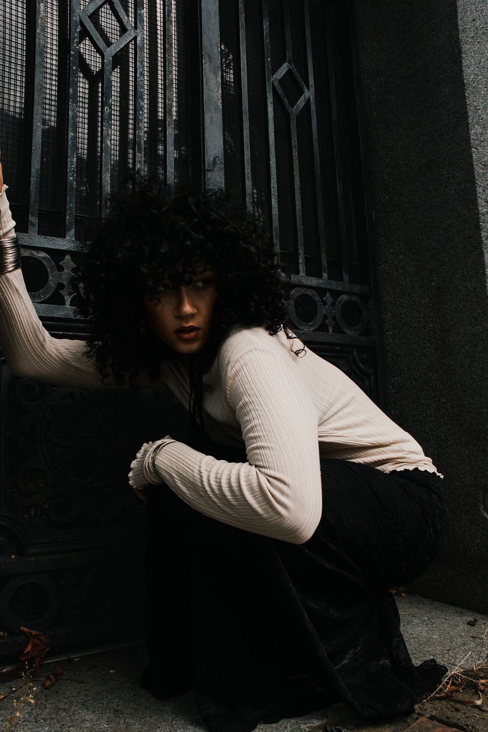Woman with voluminous curly black hair crouching beside an ornate black iron gate, wearing a cream ribbed top and long dark skirt.