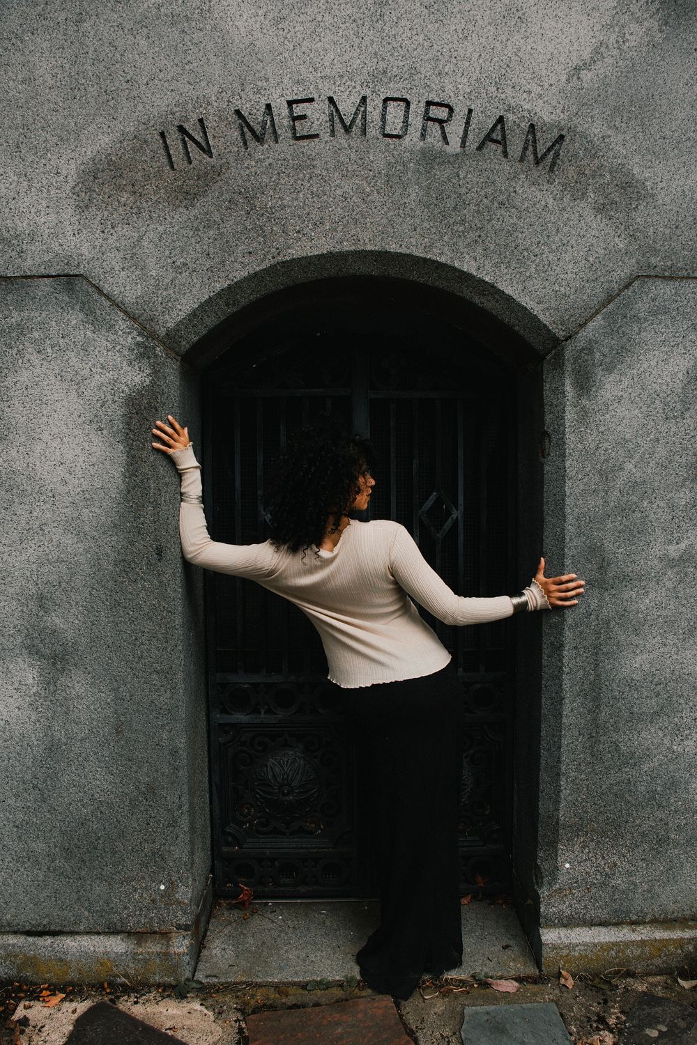 Woman in a cream top and black pants stretches her arms against a stone mausoleum entrance engraved with In Memoriam above an iron gate.