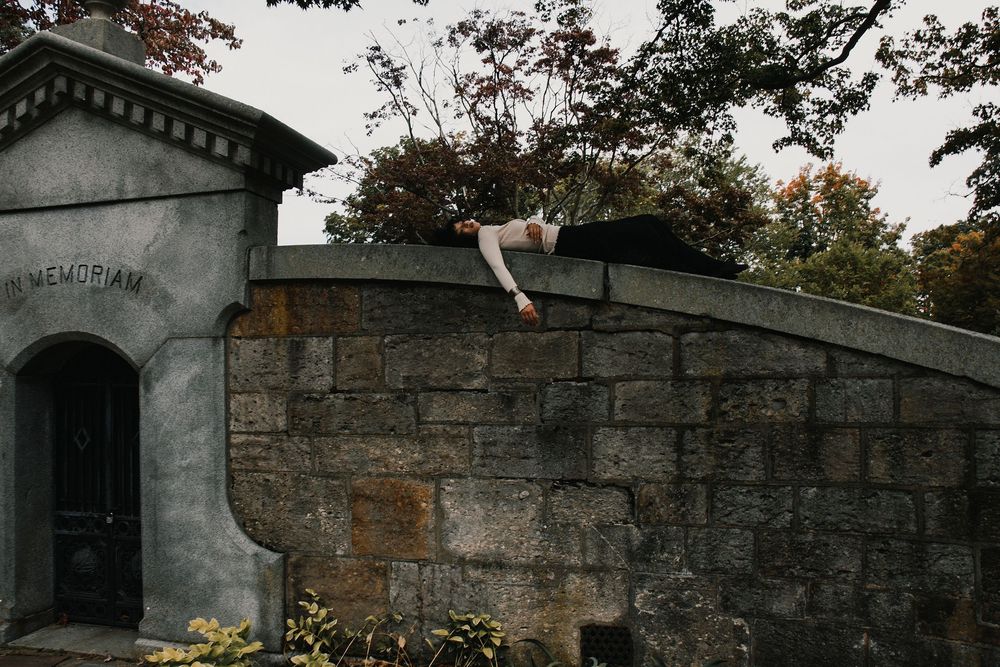 Person lying on top of a weathered stone mausoleum wall with one arm dangling over the edge, beside an arched doorway marked In Memoriam.