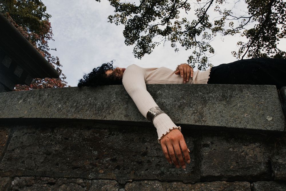 Person in a cream sweater lies draped over a weathered stone wall, one arm hanging down, with tree branches and a pale sky above.