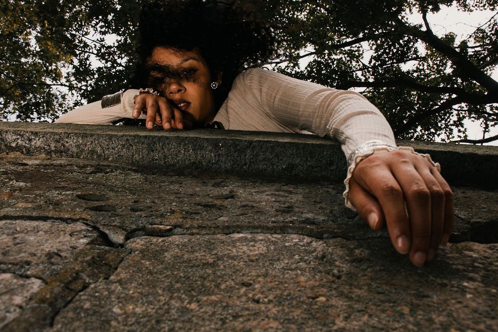 Woman with dark curly hair peers over the edge of a weathered stone wall, her hands gripping the ledge beneath leafy trees.