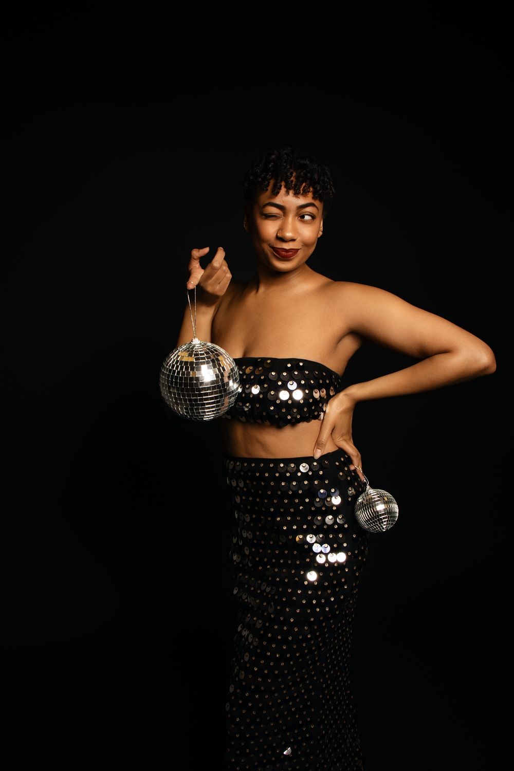 Model in a strapless sequined dress poses with a silver disco ball against dark backdrop.