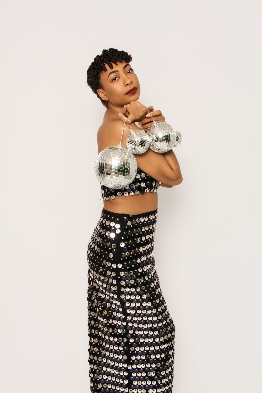 Model in sequined dress poses playfully with disco ball on white background.