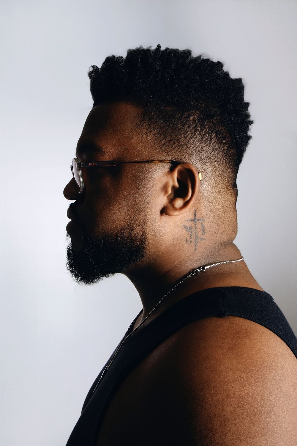 Side profile portrait of person with fade haircut and beard wearing glasses against grey background.