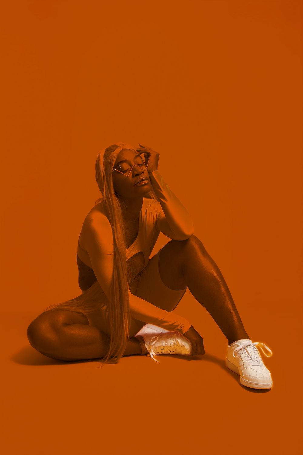 A woman in athletic wear and sneakers sits on an orange studio floor, bathed in a monochromatic orange light.