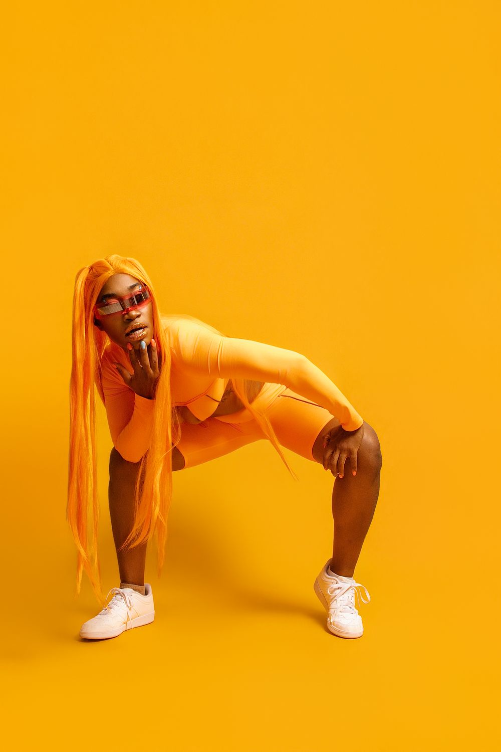 Black woman in orange bodysuit and long orange wig posing confidently against a vibrant yellow background.