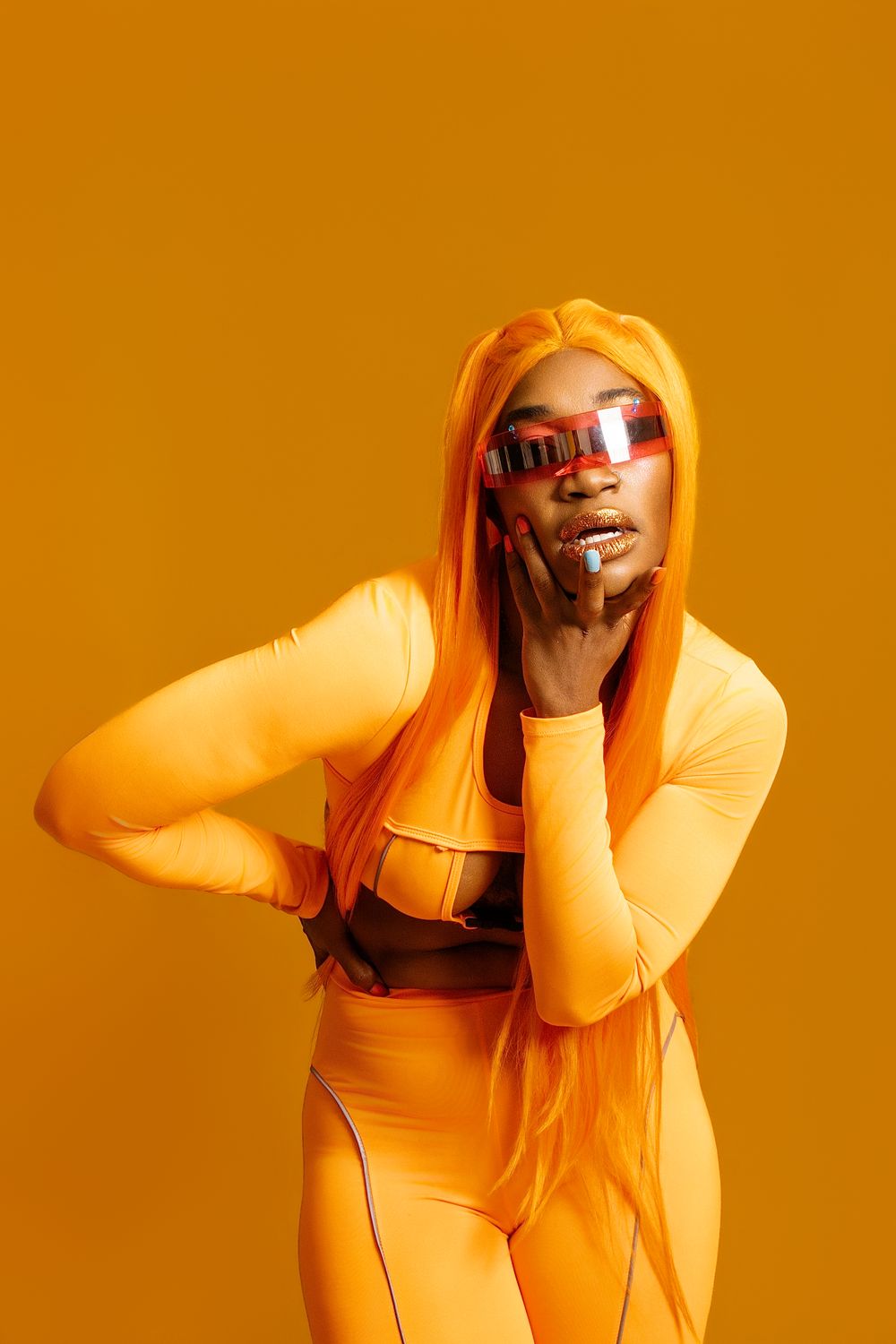 Black woman in orange outfit and wig with futuristic mirrored sunglasses posing on yellow background.