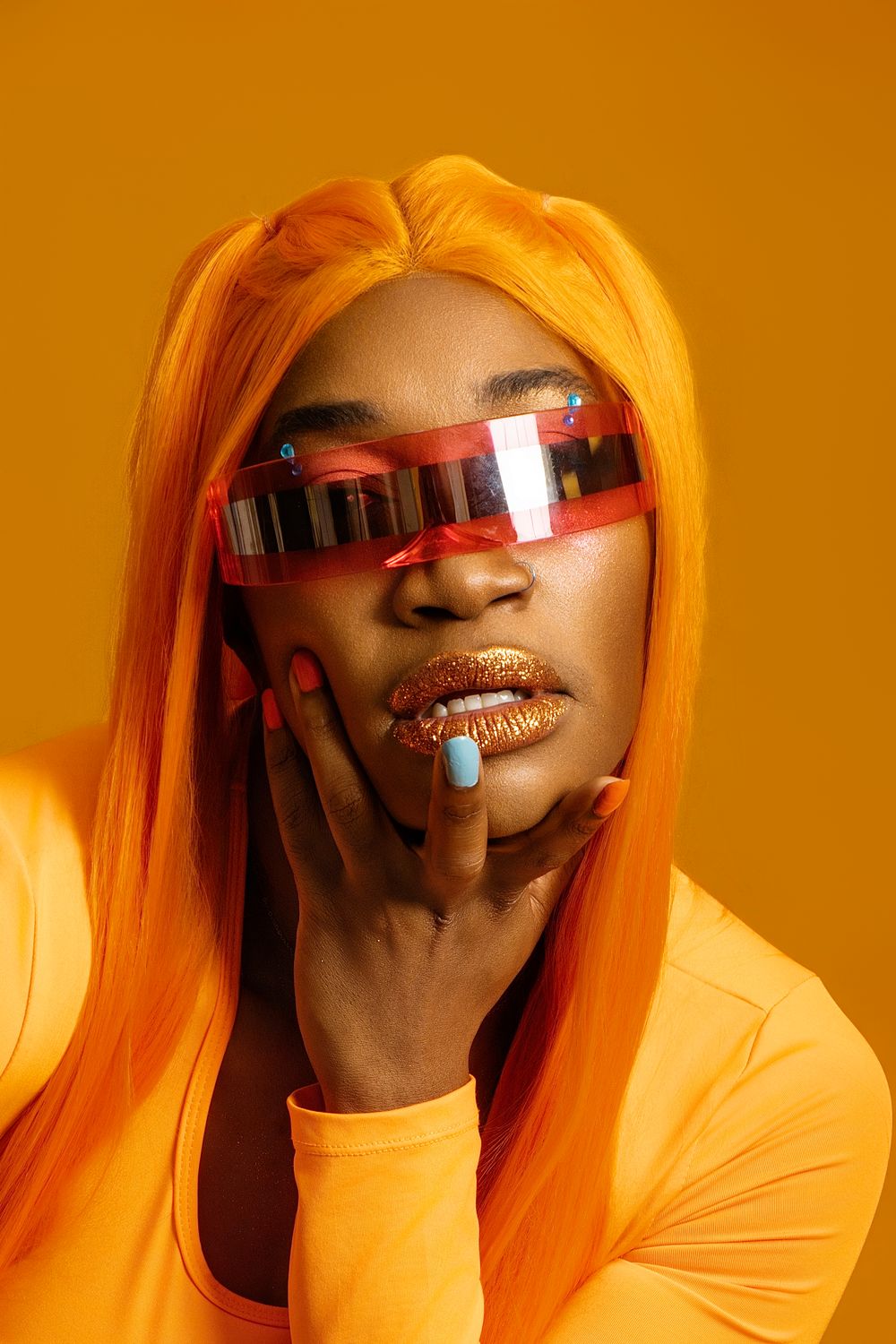 Woman in orange wig, futuristic red sunglasses, gold lips, and orange outfit poses against yellow background.