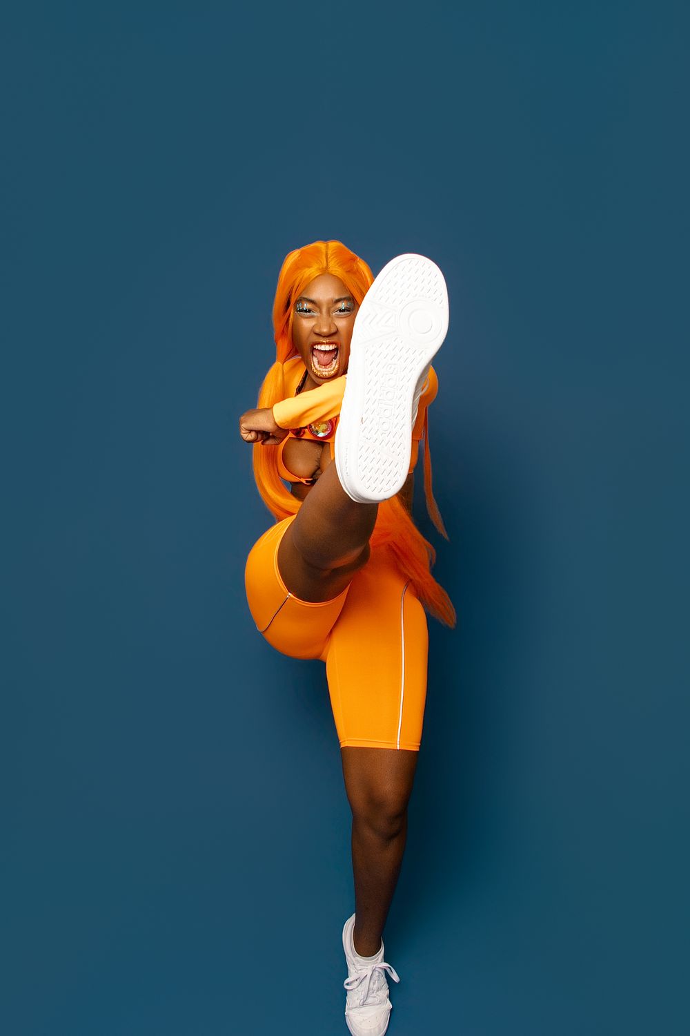 Woman in orange outfit and wig kicks leg up high against blue background, showing white sneaker sole.