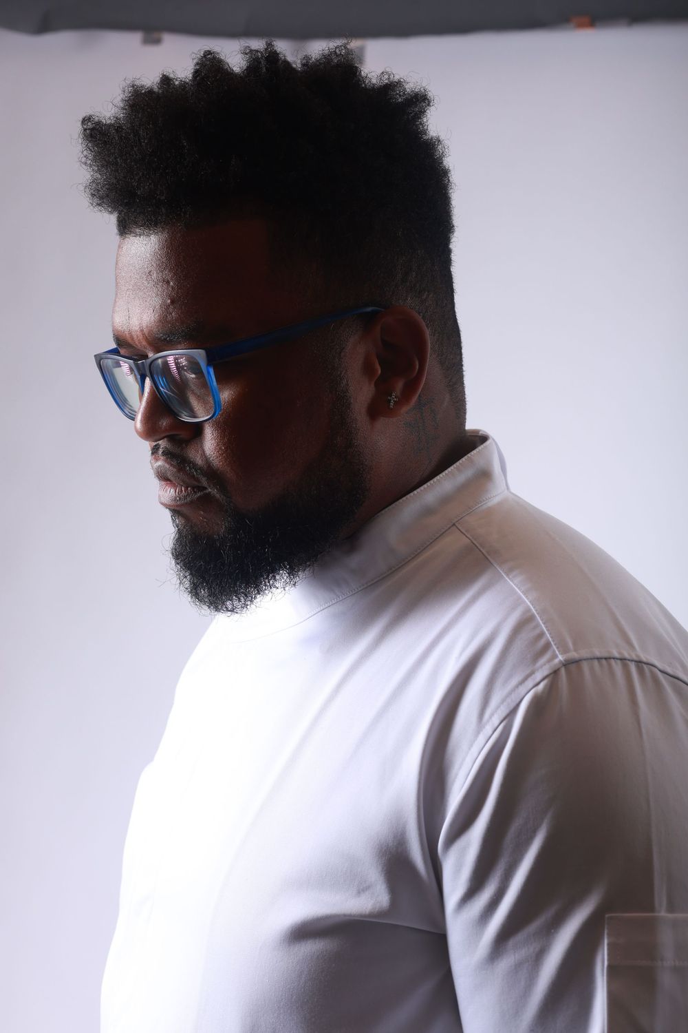 A bearded Black man with blue rectangular glasses and natural hair wearing a white turtleneck shirt poses in a studio.