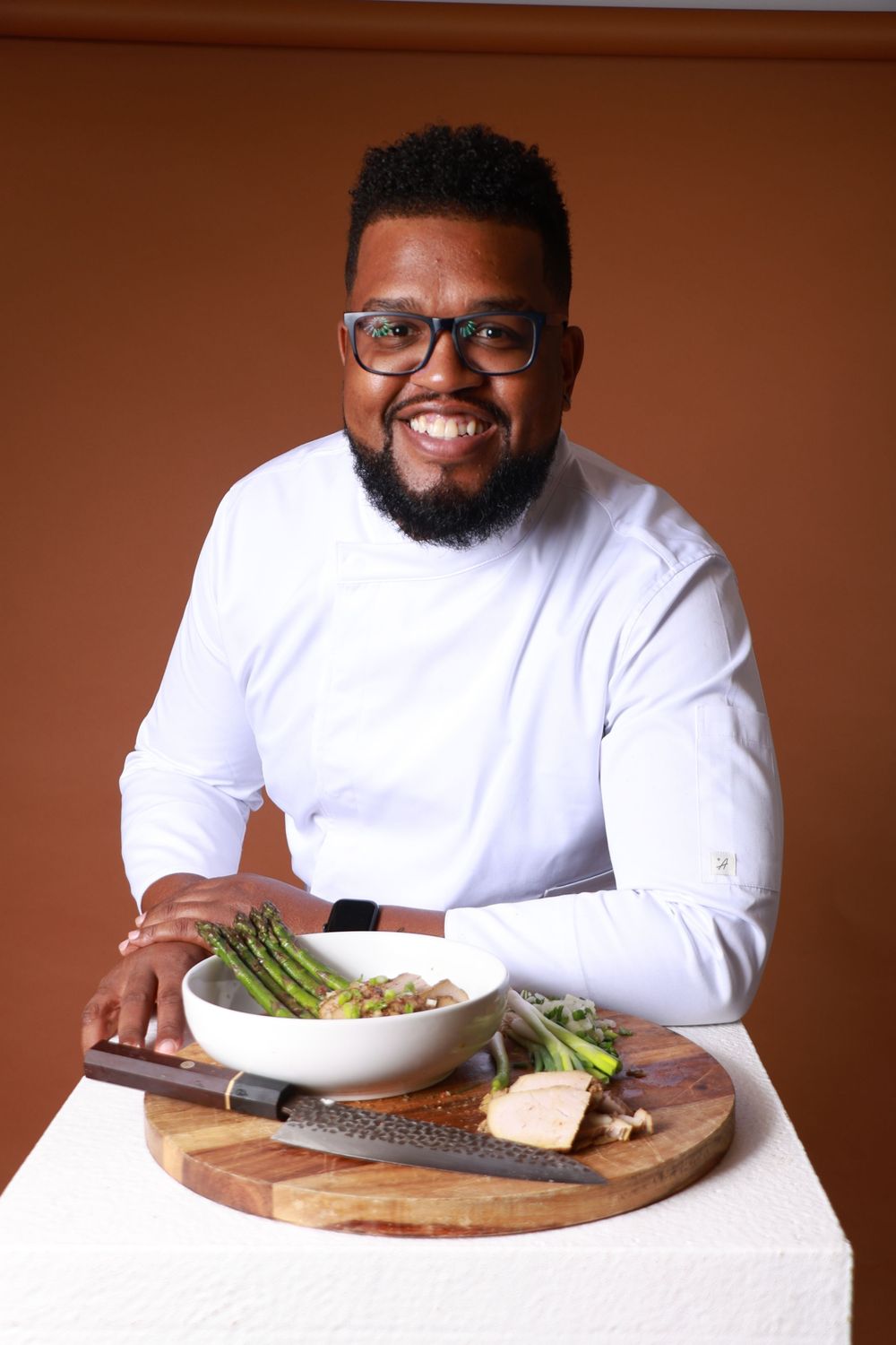 Smiling chef in white coat poses with asparagus dish on wooden board with knife.