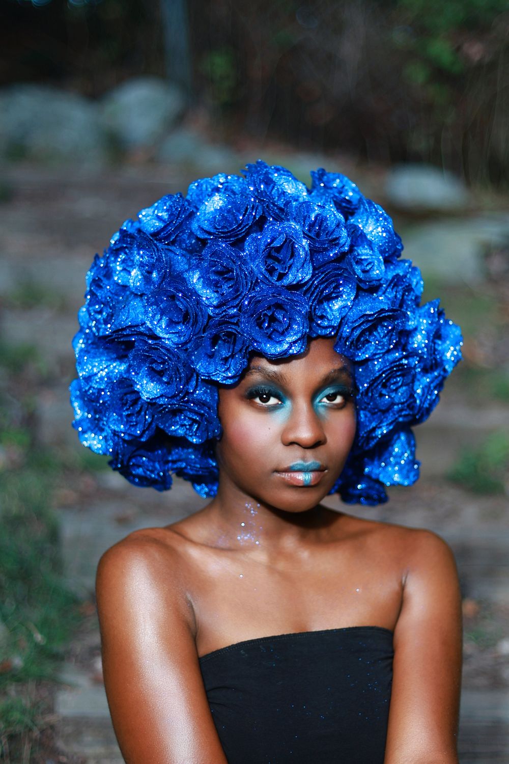 Woman wearing a sparkling blue rose-shaped headpiece with matching blue eye makeup and a black strapless top, posing outdoors near rocks.