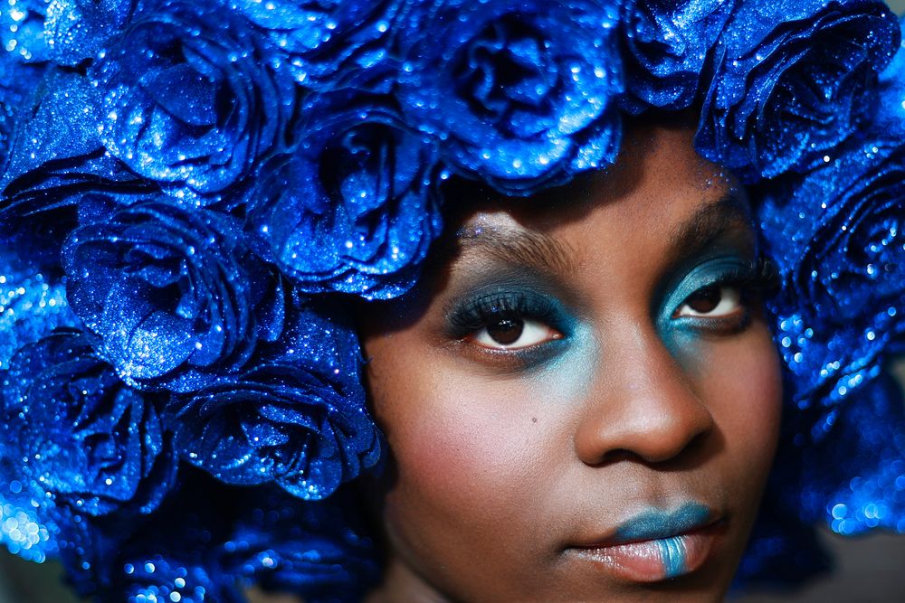 Close-up portrait of a woman wearing a voluminous headpiece of glittering royal blue roses with matching blue eyeshadow and lipstick.