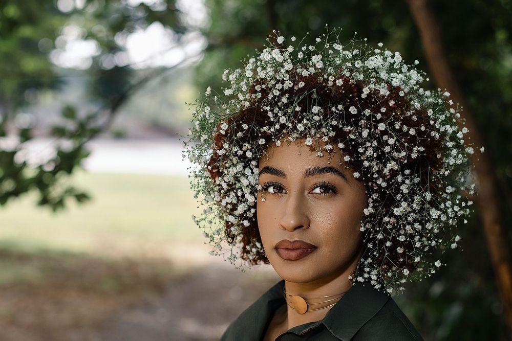 Woman with curly hair crowned in a halo of white baby's breath flowers poses outdoors in a green park, wearing a dark collared jacket.
