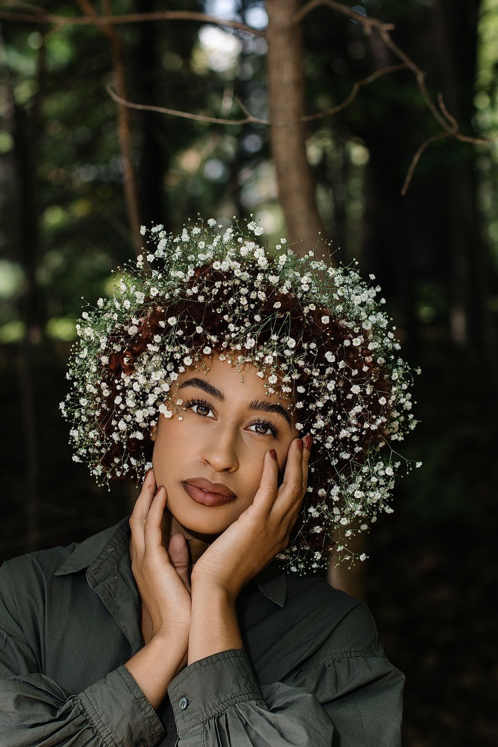 Woman in an olive green shirt poses with hands on her cheek, wearing a baby's breath flower crown in a softly lit forest.