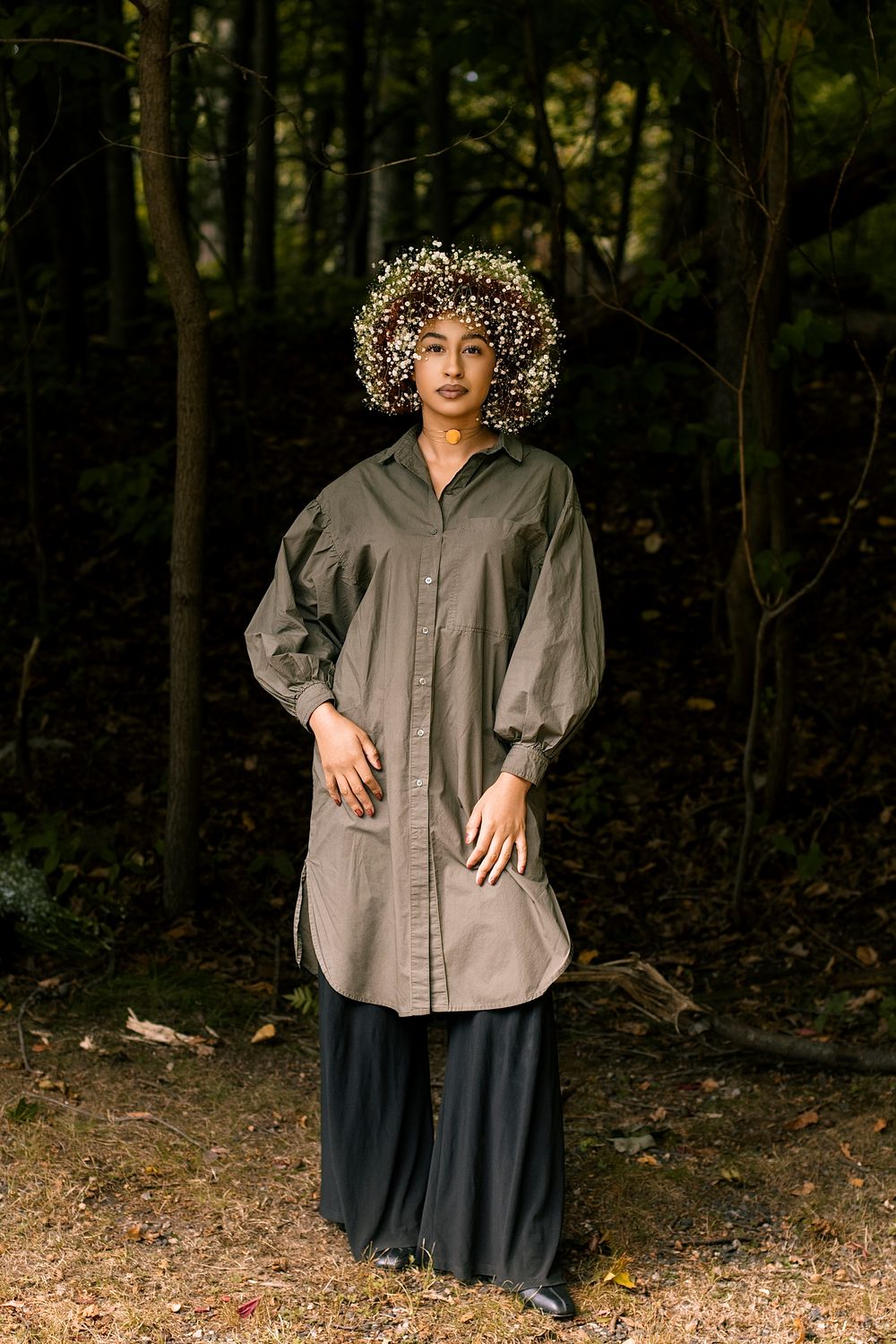 Woman in an olive oversized button-up tunic and wide-leg black pants wears a baby's breath floral crown while standing in a dark forest.
