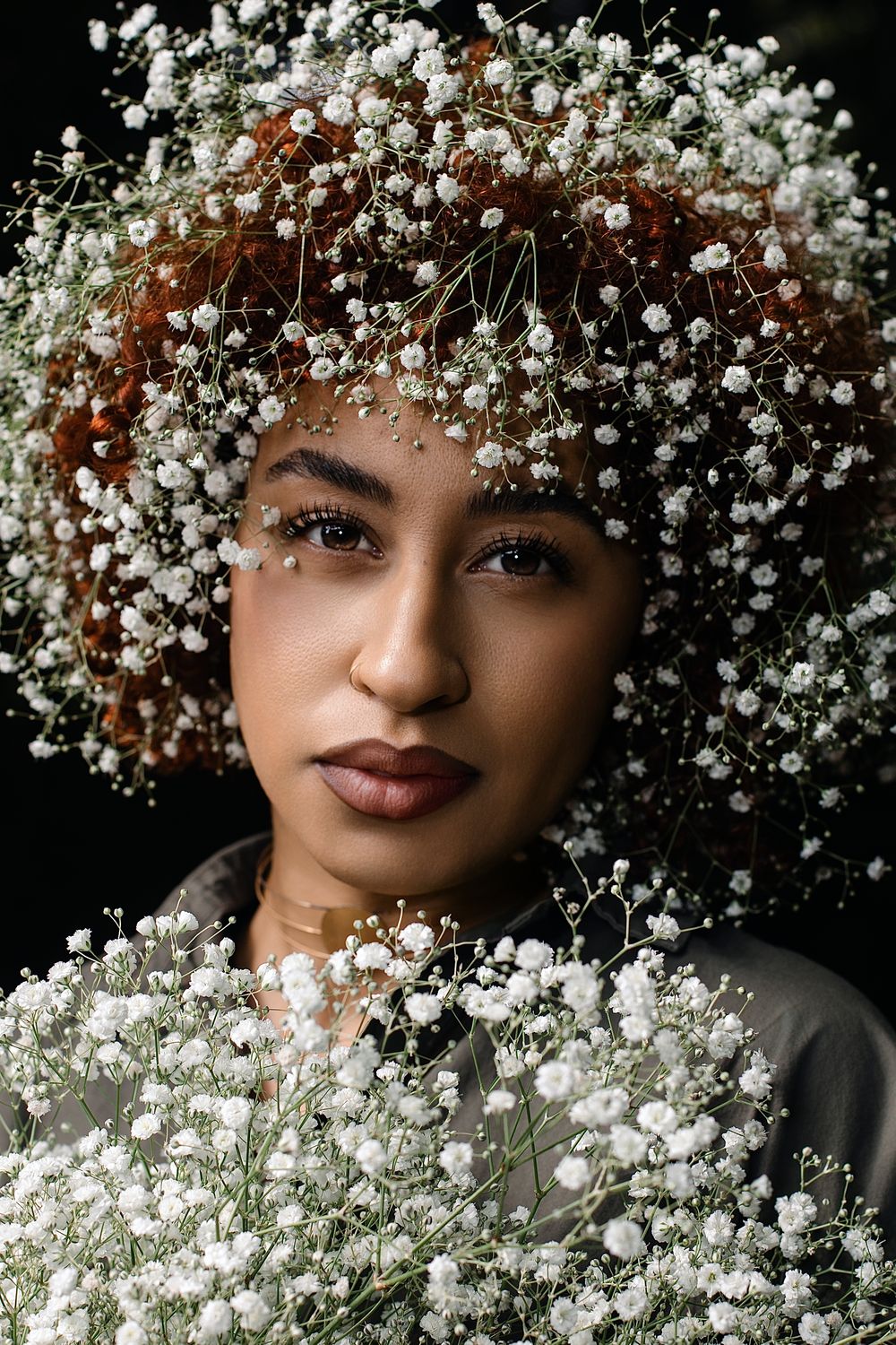 Portrait of a woman with auburn curly hair covered in delicate baby's breath flowers, holding a bouquet against a dark background.