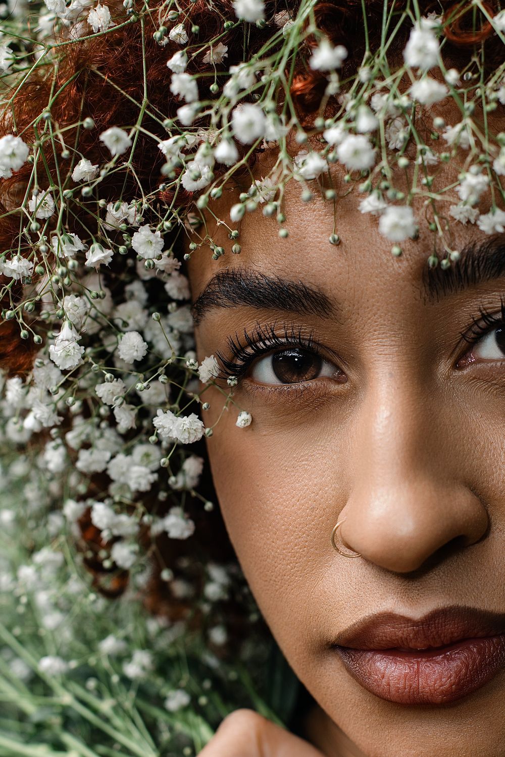 Close-up portrait of a woman with curly auburn hair adorned with delicate white baby's breath flowers, partially framing her face.