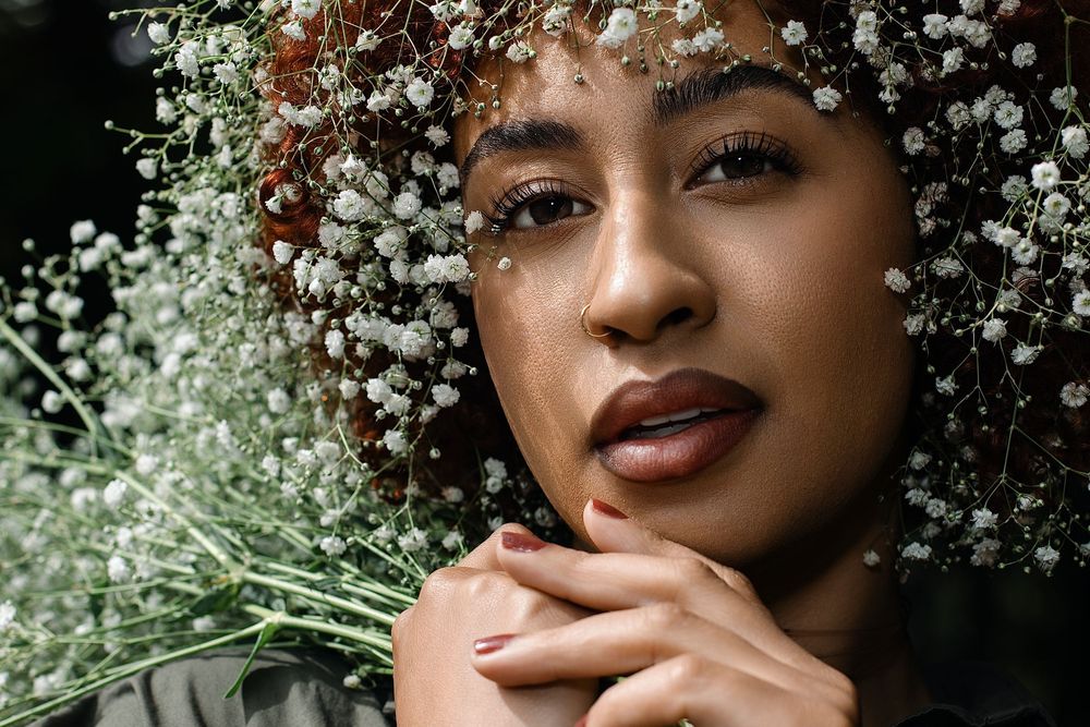 Portrait of a woman with curly hair adorned with delicate white baby's breath flowers, hands gently posed beneath her chin.