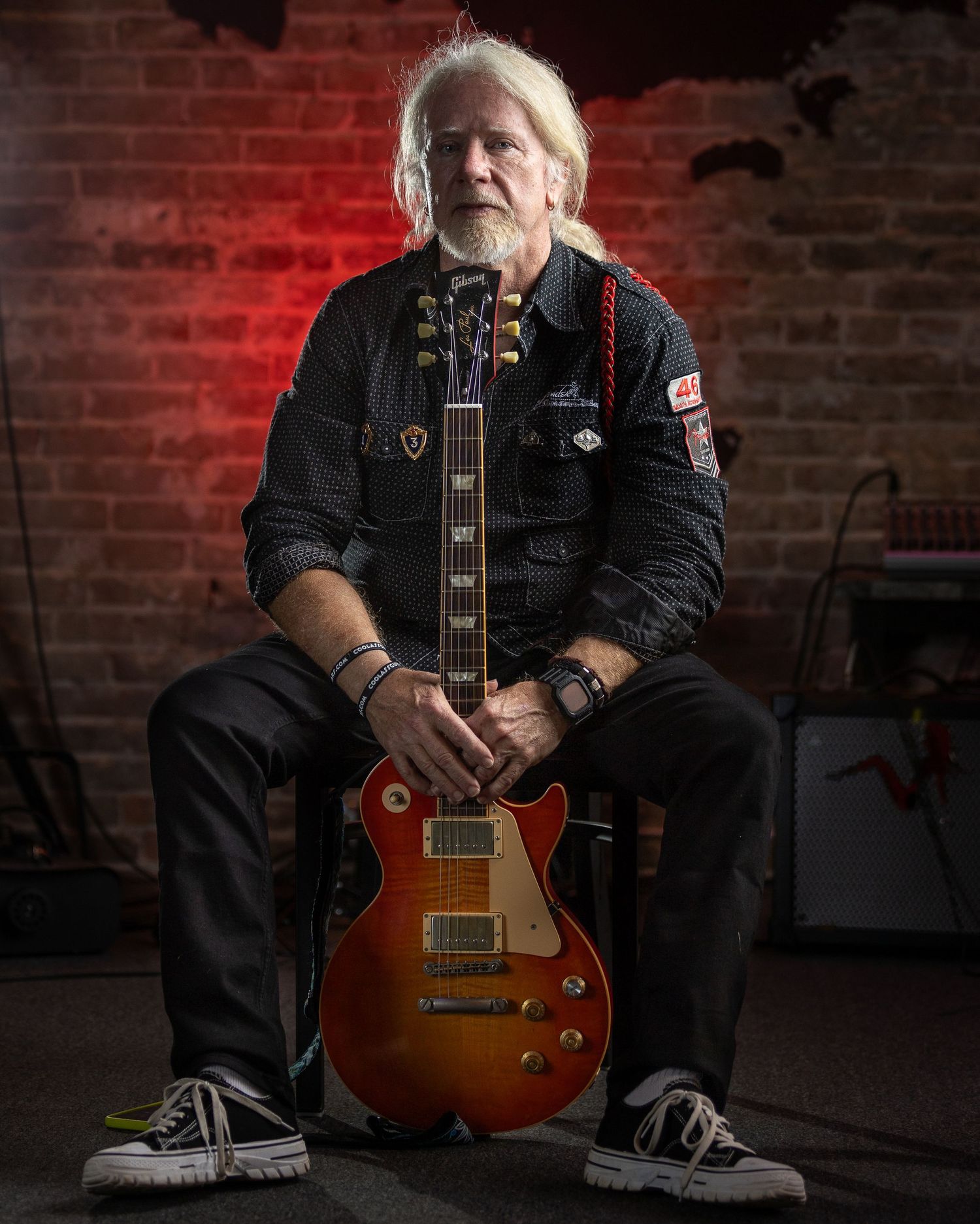 Professional studio portrait of a musician in black denim holding a vintage sunburst Les Paul guitar against a red brick wall.