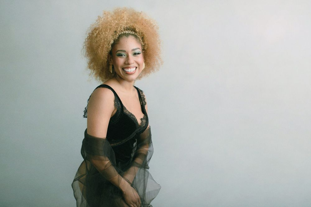 A model with curly blonde hair poses in a black dress against a light background for a professional photoshoot.