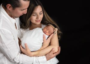 Two parents in white garments lovingly holding their sleeping newborn baby against a dark background.#newbornphotography Artesia