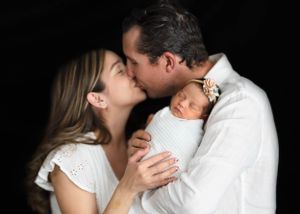 A tender family portrait against black backdrop shows parents sharing a kiss while holding their sleeping newborn baby. #newbornphotography