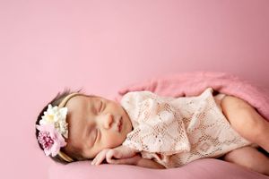 Sleeping newborn in delicate lace outfit and floral headband rests peacefully on pink background.#newbornphotography #babypictures Carlsbad