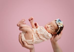 A newborn baby wearing a lace dress and floral headband is held aloft against a pink backdrop #Newbornbabyphotography #newbornphotography