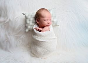 Peaceful newborn baby swaddled in white blanket sleeping on checkered pillow against soft white furry background. Newborn Photographer NM