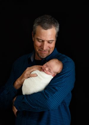 Tender moment captured in dark lighting shows someone in a blue plaid shirt cradling a sleeping newborn wrapped in white blanket. New Mexico