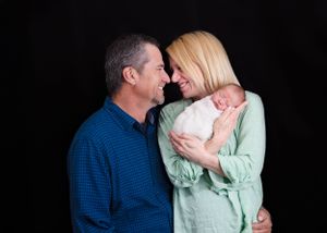 A loving couple in plaid shirt and mint green sweater share a tender moment while holding their newborn baby against black backdrop. NM