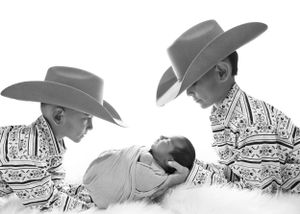A black and white photograph shows two siblings in cowboy hats and patterned shirts looking down at a small baby in between them. Newborn