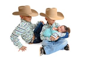 Siblings in western attire with cowboy hats and matching shirts hold newborn brother swaddled in blue. Baby photographer Carlsbad New Mexico