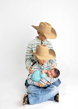 Siblings in western attire with cowboy hats and matching shirts hold newborn brother swaddled in blue. Baby photographer Carlsbad New Mexico