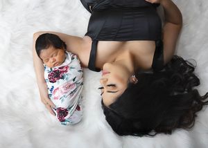 A baby wrapped in floral swaddle blanket lies on white fabric next to someone wearing black. #NewbornFamilyPictures #NewbornPhotographer